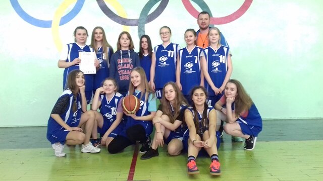 Zdjęcie przedstawia drużynę dziewcząt w strojach sportowych wraz z trenerem. Zawodniczki prezentują dyplom mistrza zawodów rejonowych w minikoszykówce dziewcząt.